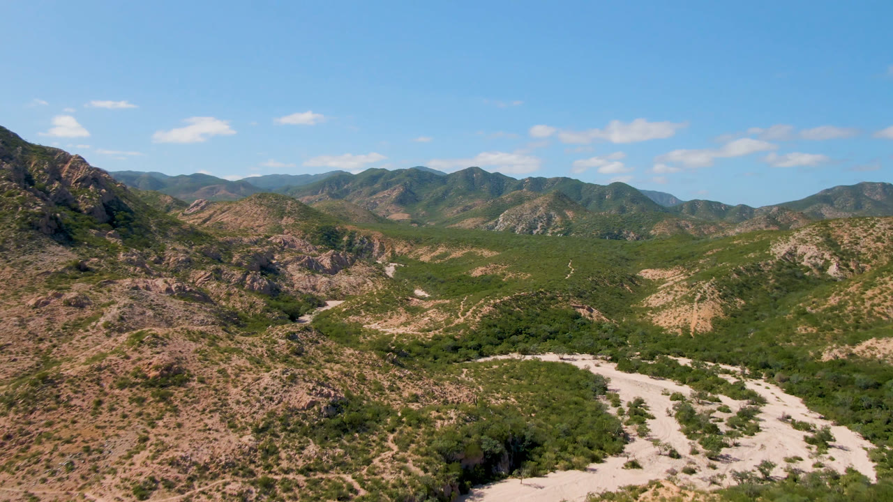 Opening aerial frame over Cabo Pulmo, Baja California Sur, with rocky desert hills, green scrub in the valley, pale winding tracks, distant ridges, and no visible people.