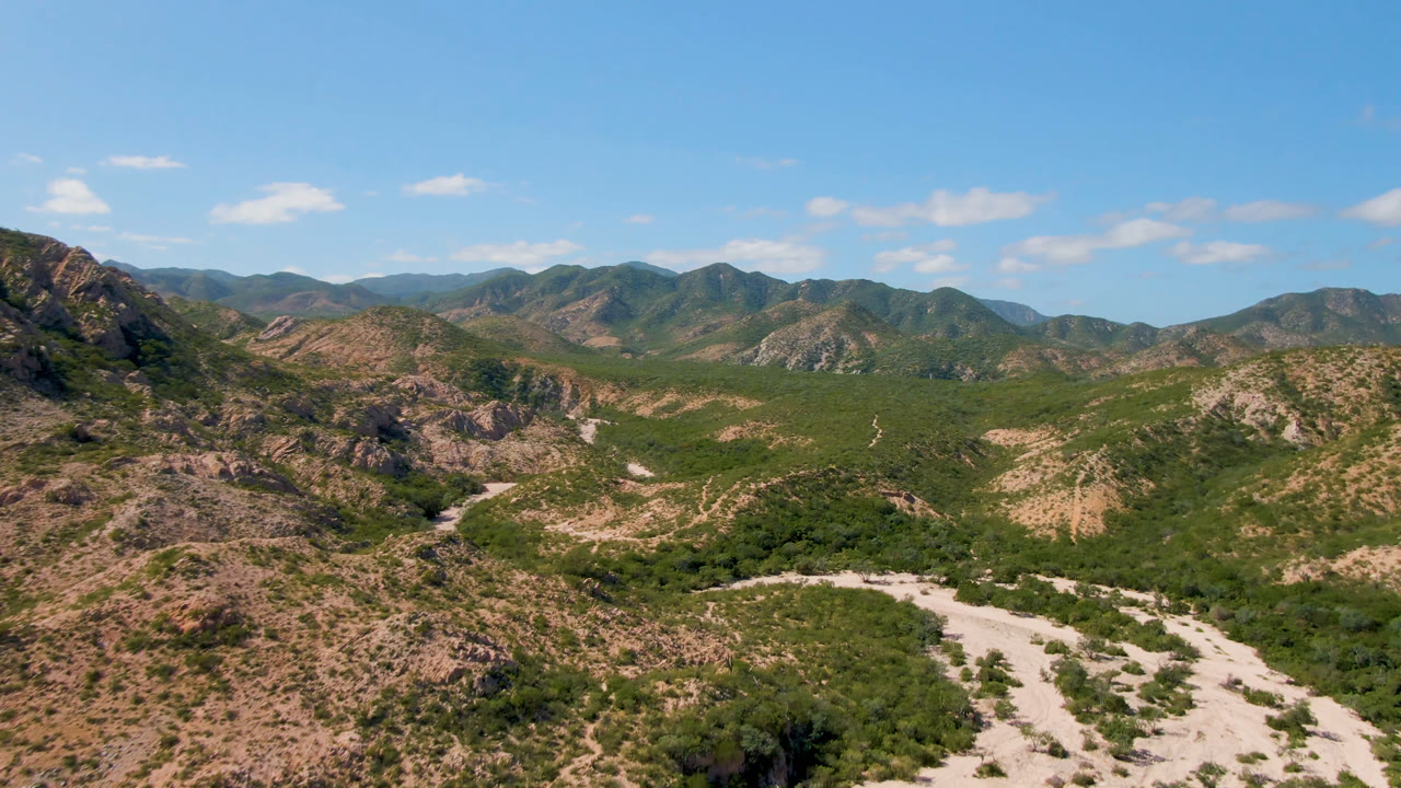 Second aerial frame in Cabo Pulmo showing a darker rocky slope at left, scrub-filled lowland, a pale road or dry wash curving through the foreground, and no visible people.