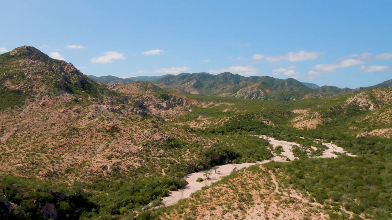 Elevated Cabo Pulmo landscape with a central rocky peak, scrub-covered slopes, pale dry riverbed channels in the valley, distant mountain ridges, and no visible people.