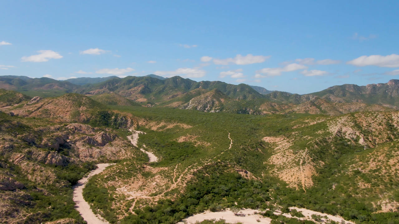 Wide desert mountain valley with rocky hills, green vegetation, and winding road cuts.