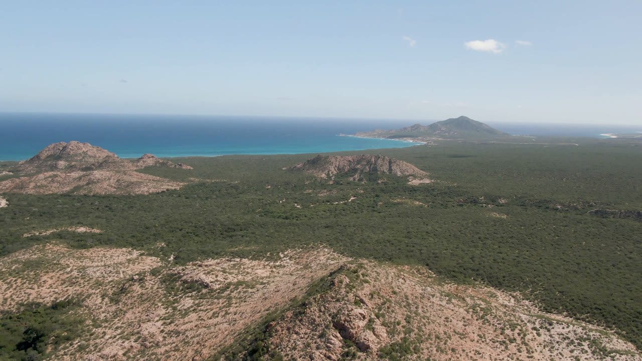 Wide aerial view of rocky scrubland leading to blue ocean under clear sky.
