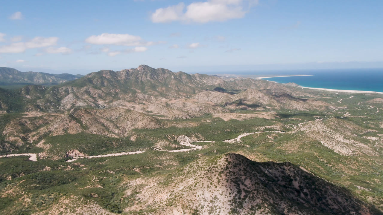 Wide aerial frame over Cabo Pulmo arid mountains, green desert vegetation, a pale road, distant blue ocean, small clouds, and no visible people.
