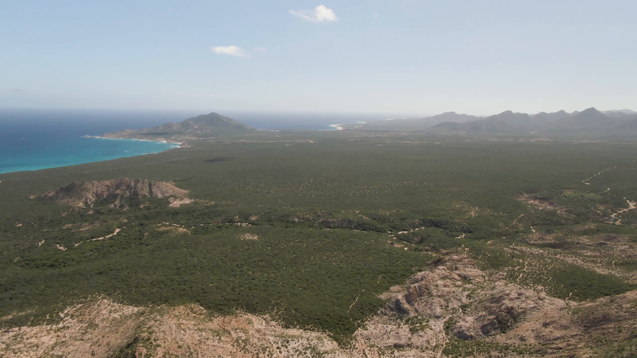 Coastal aerial view with ocean left, low rocky hills, green plain, and mountains beyond