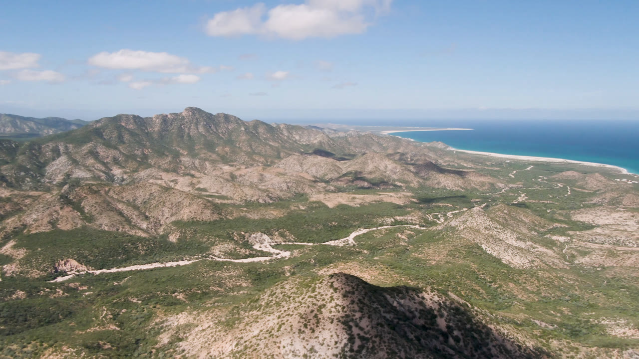 Aerial frame over Cabo Pulmo with a rounded foreground hill, rocky mountain ridges, a winding pale road through desert vegetation, blue ocean on the right horizon, and no visible people.