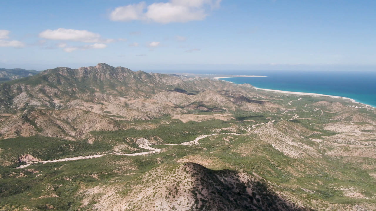 A high aerial view in Cabo Pulmo shows tan mountain slopes, green desert patches, a pale road curving through the valley, a bright coastline to the right, and no visible people.