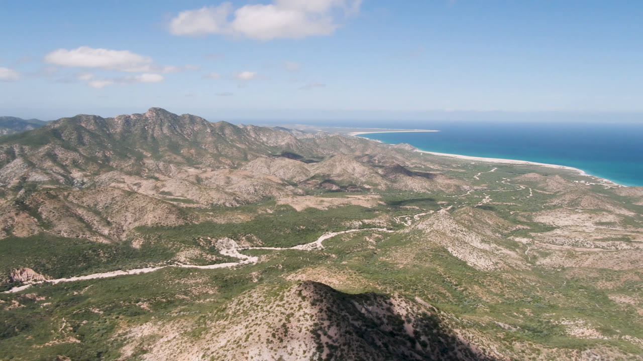 Wide aerial landscape of rugged mountains, scrubland roads, and blue sea beyond.