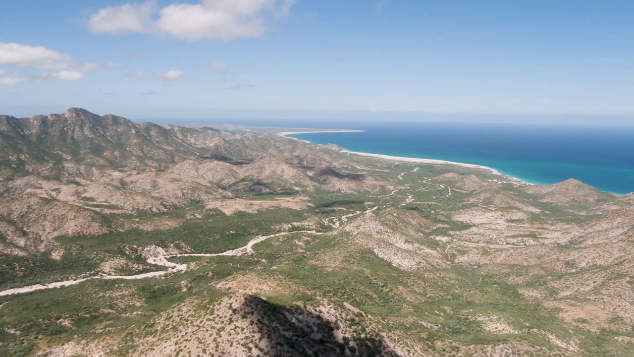 Aerial frame from Cabo Pulmo showing rocky tan hills, green scrub, a pale winding road, turquoise ocean, distant beach line, scattered clouds, and no visible people.