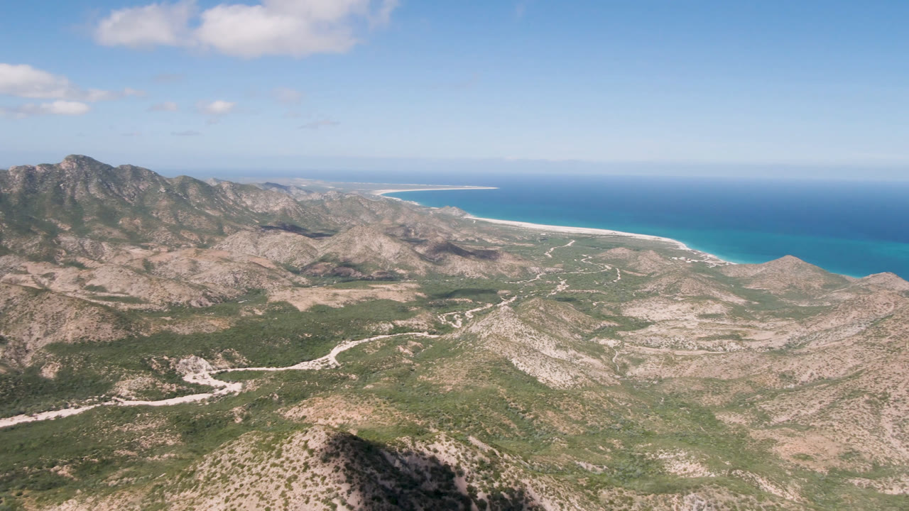 Second frame from Cabo Pulmo holds a wide aerial view of arid mountain ridges, road curves across the valley floor, blue ocean bands, small clouds, and no visible people.