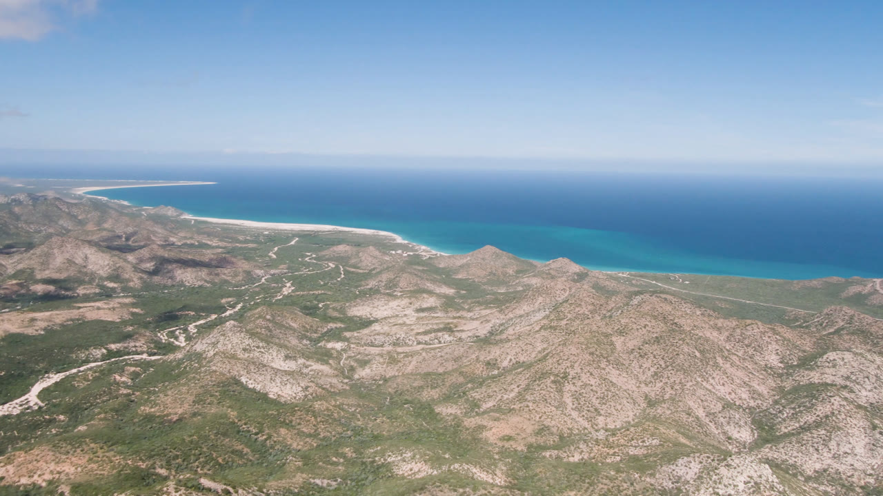 Wide aerial view of rocky coastal mountains, blue ocean, and pale sky.