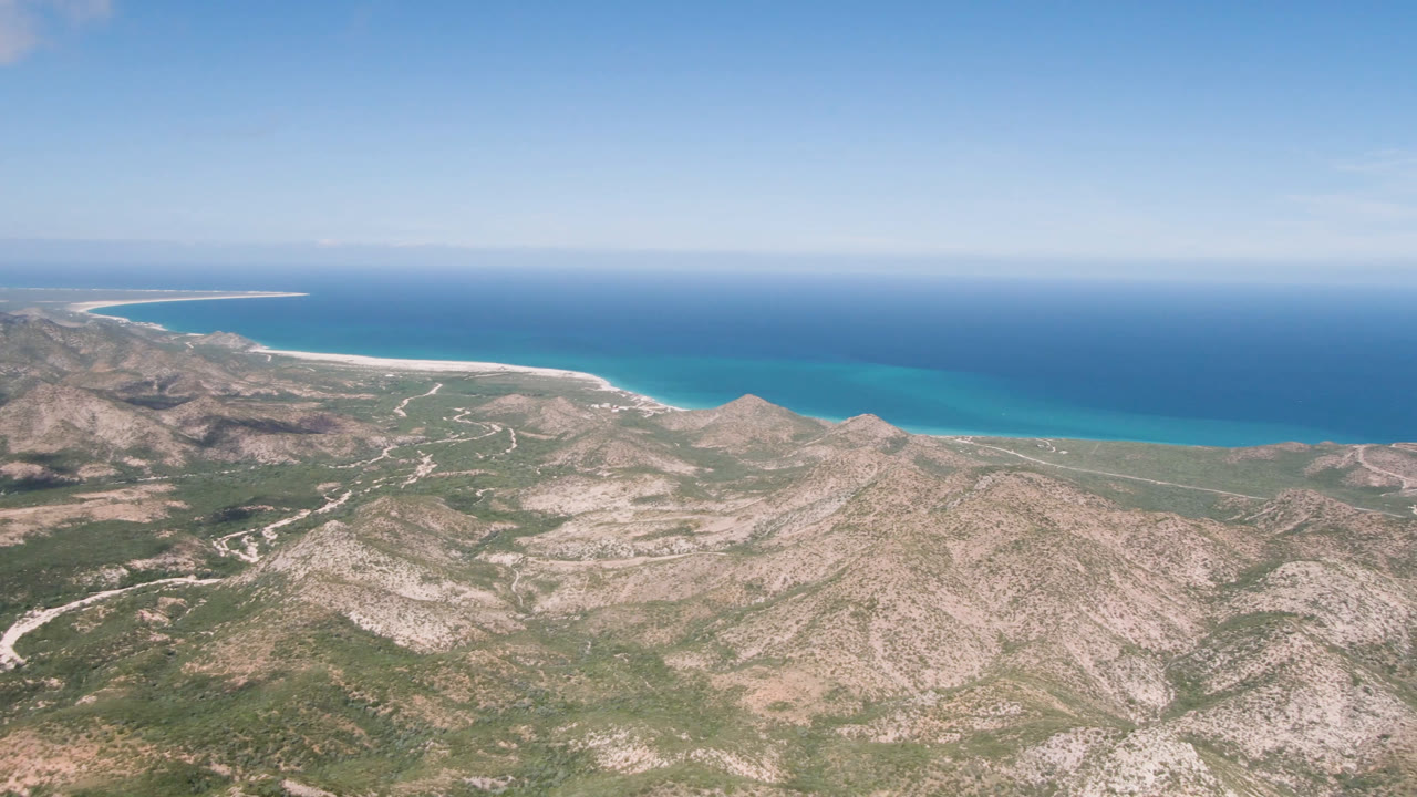 Aerial frame of Cabo Pulmo showing rugged tan hills, scattered green vegetation, a narrow beach edge, and open blue water with no visible people.
