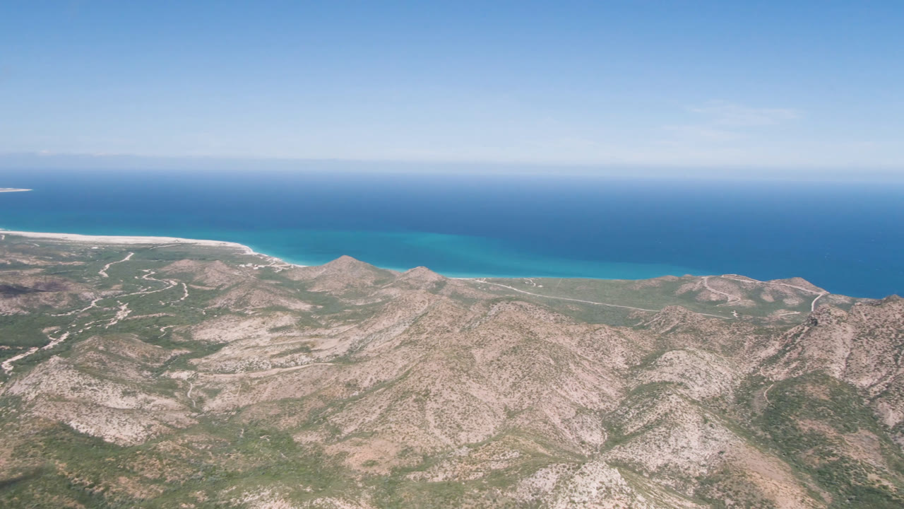 Wide aerial frame over Cabo Pulmo, Mexico, with pale rocky ridges, sparse green desert cover, turquoise shoreline, deep blue ocean, clear sky, and no visible people.