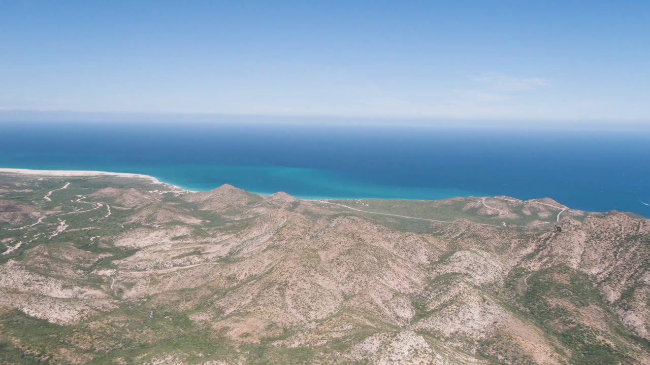 Aerial view in Cabo Pulmo, Mexico, showing dry ridgelines across the lower frame, a thin pale beach edge, blue-green shallows, darker open water, and no visible people.