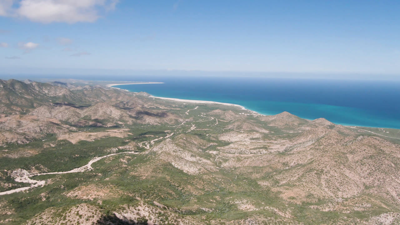 Aerial view over Cabo Pulmo rocky mountain terrain with winding pale roads, turquoise shoreline, blue ocean horizon, scattered clouds, and no visible people.
