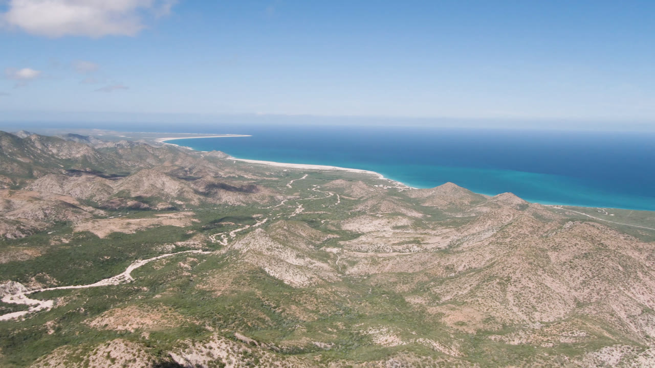 High aerial frame of Cabo Pulmo arid hills and green scrub, with pale tracks crossing the land toward a bright blue coastal waterline and no visible people.