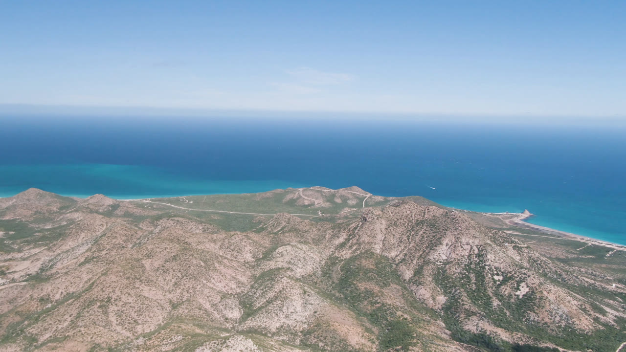 Wide aerial frame over Cabo Pulmo, Baja California Sur, with rocky mountain slopes below, turquoise shoreline water beyond, deep blue ocean at the horizon, and no visible people.