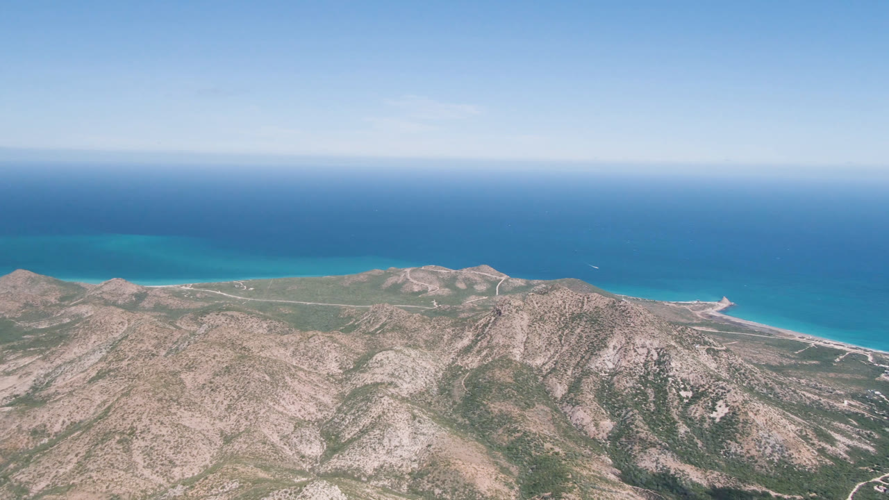 Aerial view in Cabo Pulmo, Baja California Sur, showing a low coastal plain between rugged hills and a band of turquoise ocean, with no visible people.