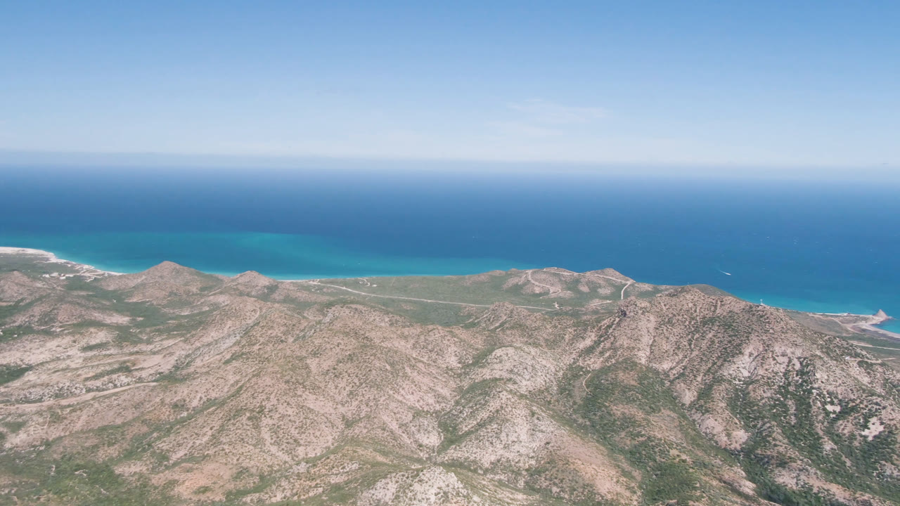 At 0:01, the Cabo Pulmo coastline remains wide in frame, with pale ridges, green scrub patches, a thin beach edge, blue ocean, and no visible people.