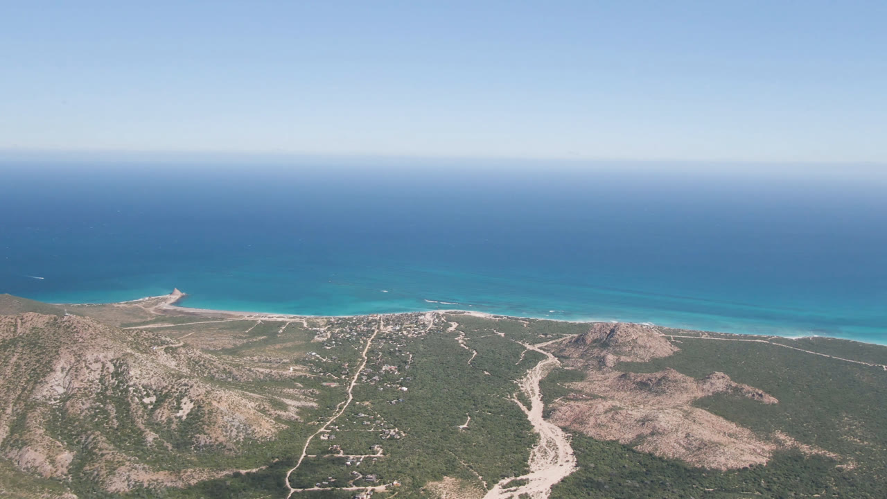 High aerial view over Cabo Pulmo, Mexico, with scrubby desert hills, pale dirt roads, a thin beach, turquoise shoreline water, deep blue ocean, clear sky, and no visible people.
