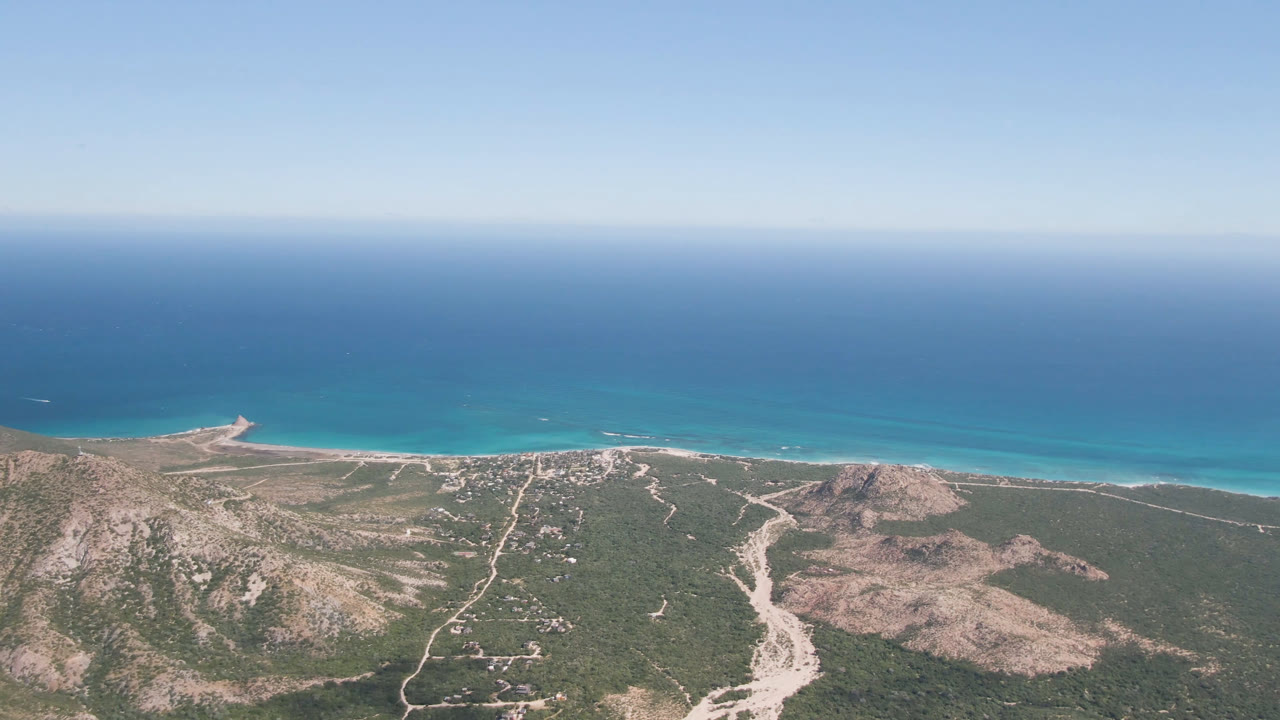 Aerial frame from Cabo Pulmo, Mexico, showing the coastline slightly farther along, with a pale road network across green-brown scrub, rocky hills, turquoise shallows, open ocean, and no visible people.