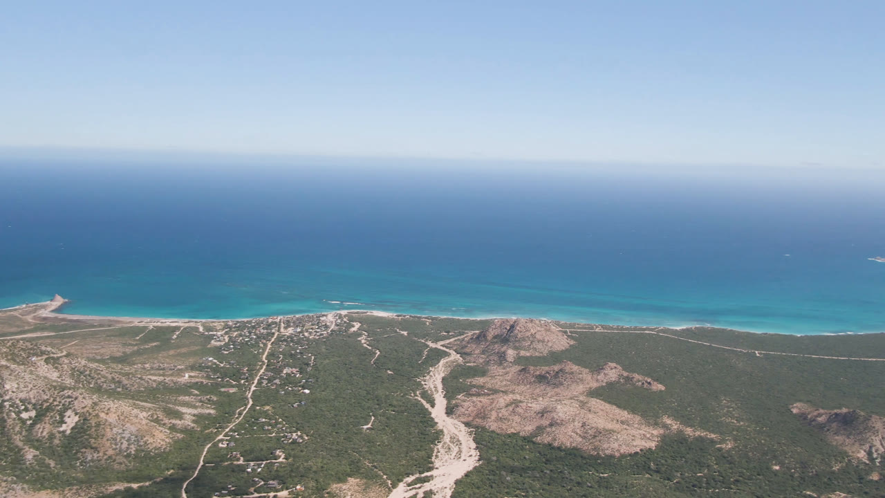 Wide aerial frame over Cabo Pulmo, Baja California Sur with deep blue ocean, turquoise shallows, a pale beach strip, desert roads, rocky hills, and no visible people.