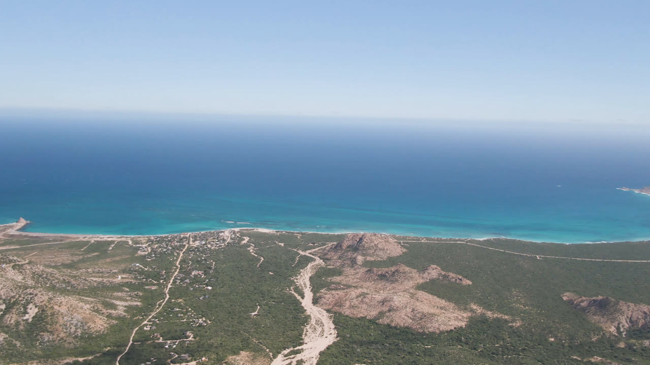 Aerial view over Cabo Pulmo with the ocean horizon across the top, turquoise water near shore, dirt tracks through scrubland, low rocky hills, and no visible people.