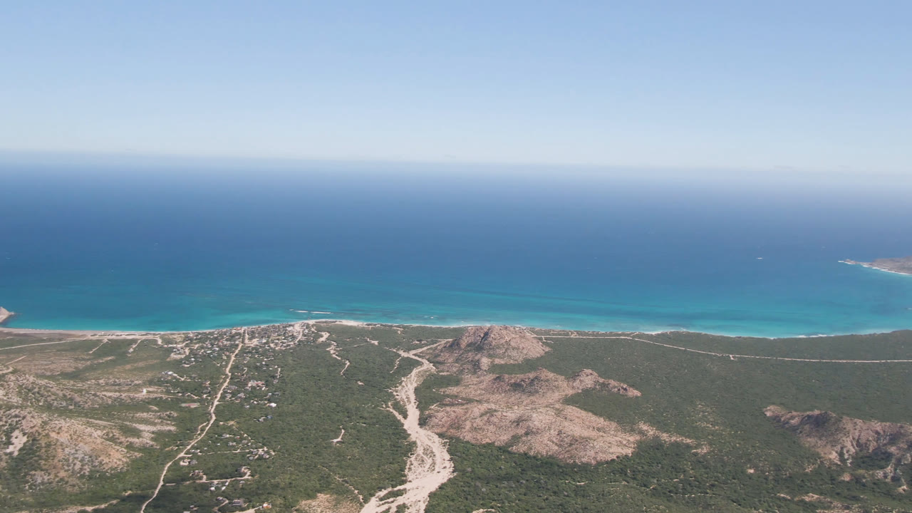 Aerial coastline with broad blue water, beach edge, dirt roads, and hills.