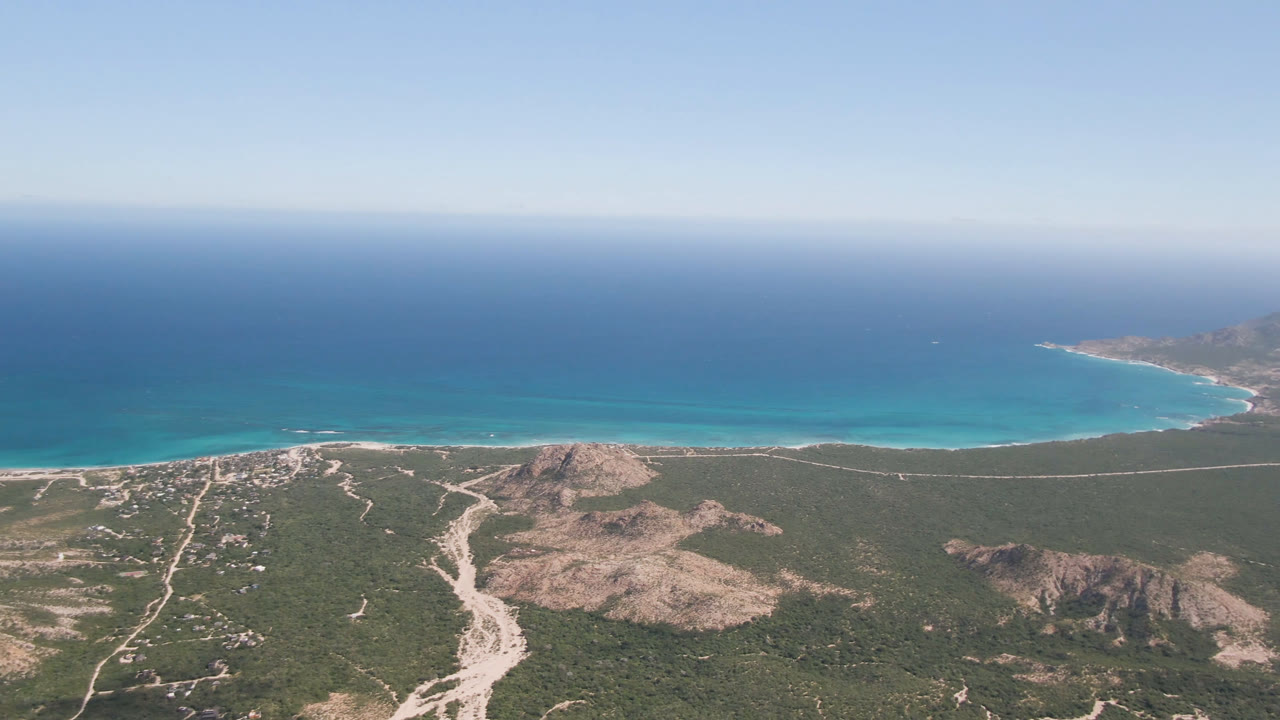 Aerial view over Cabo Pulmo, Baja California Sur, with scrub-covered desert hills, pale dirt tracks, a narrow beach, blue ocean, and no visible people.