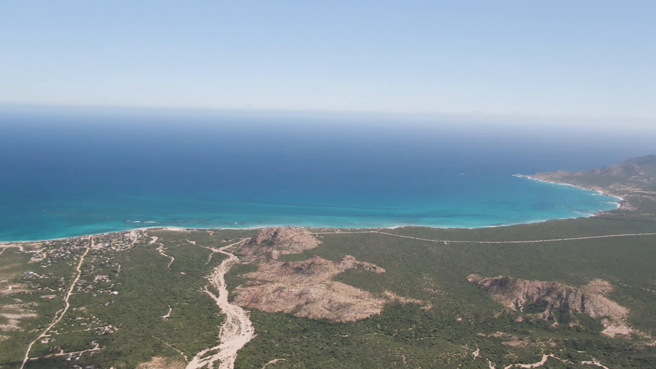 Wide aerial view of Cabo Pulmo coastline with rocky desert rises in the foreground, branching dirt roads, turquoise water along the beach, and no visible people.