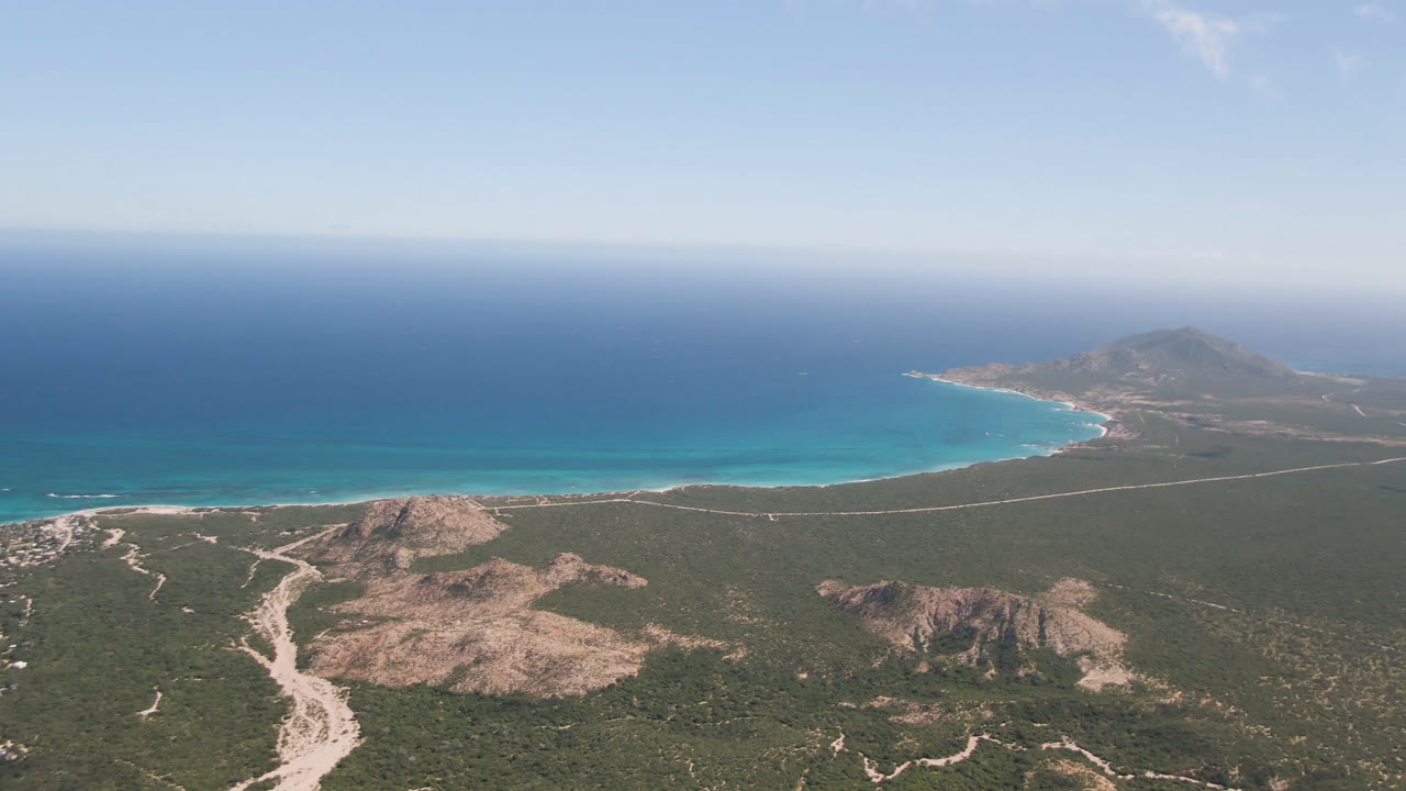 High aerial view over Cabo Pulmo, Baja California Sur, with rocky scrubland in the foreground, turquoise shallows along a curved beach, deep blue ocean beyond, and no visible people.