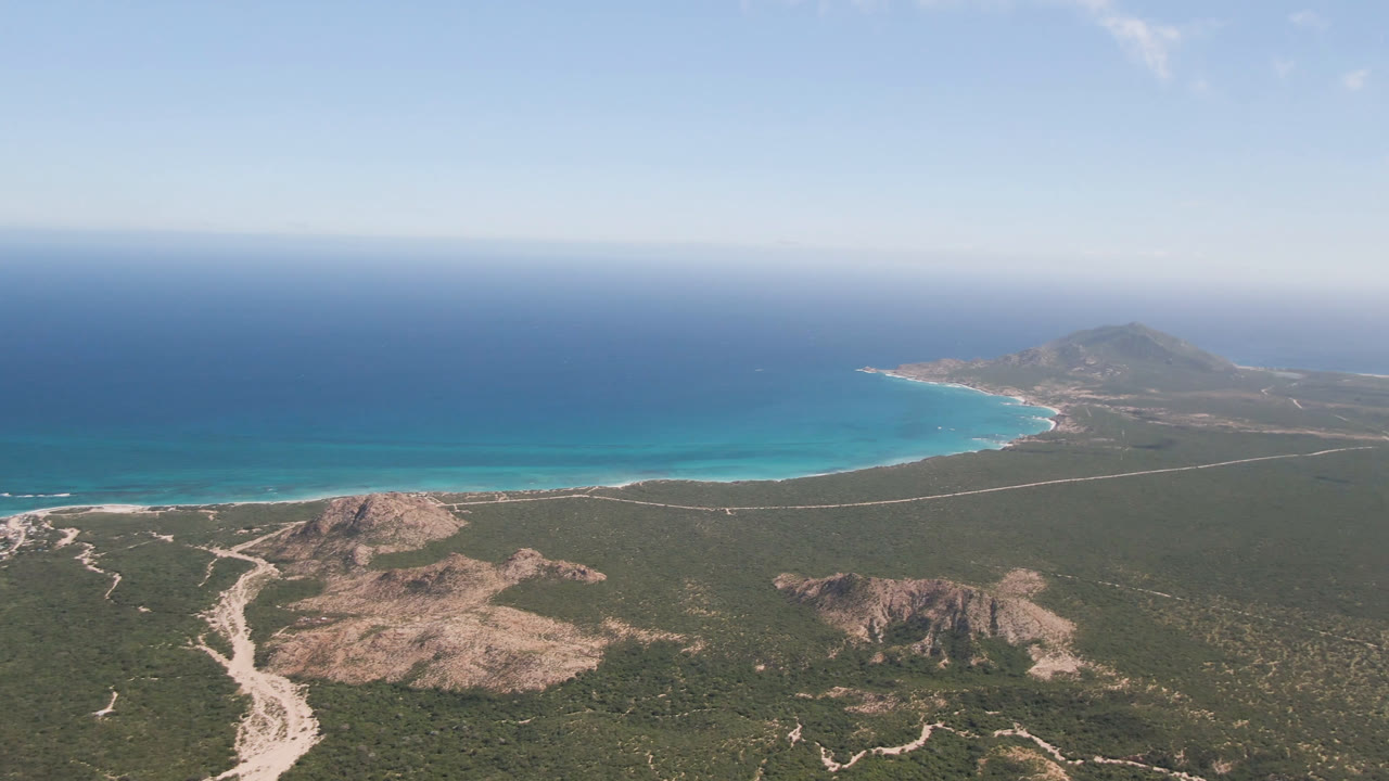 Aerial frame over Cabo Pulmo showing pale dirt tracks through green scrubland below, a bright turquoise shoreline in the midground, a distant headland to the right, and no visible people.