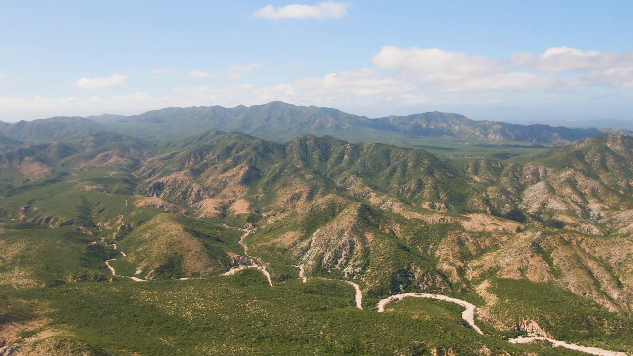 Aerial view over Cabo Pulmo, Baja California Sur, with scrub-covered ridges, pale winding routes in the lower frame, distant mountains, scattered clouds, and no visible people.