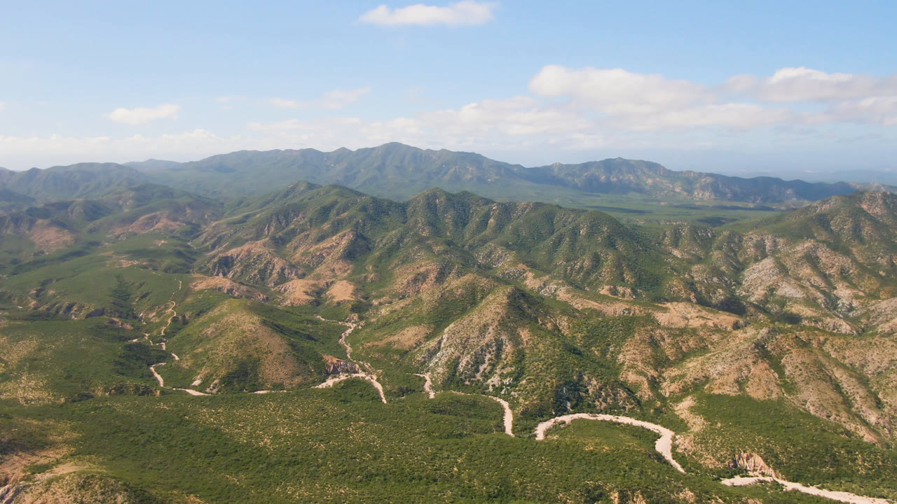 Elevated Cabo Pulmo landscape with a pale road or wash curving through the foreground, layered green and tan ridges, hazy distant hills, blue sky, and no visible people.