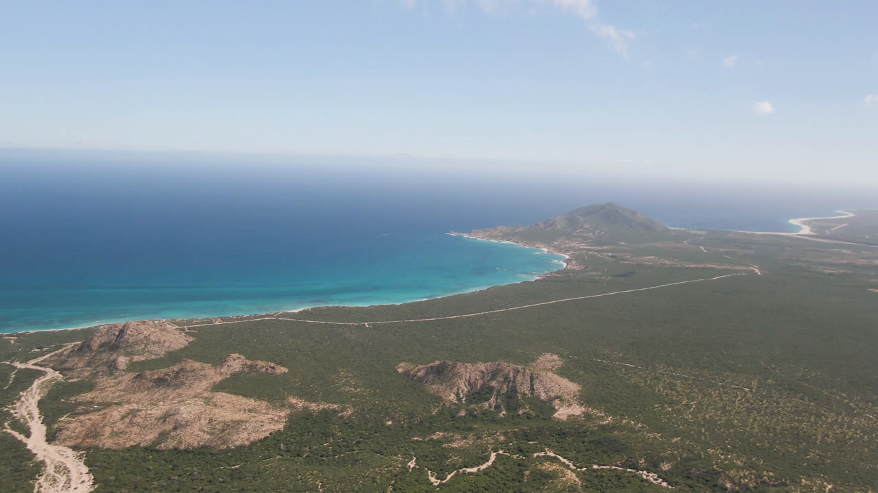 Wide aerial frame over Cabo Pulmo, Baja California Sur, with rocky scrubland in the foreground, a curved blue shoreline in the midground, open ocean beyond, and no visible people.