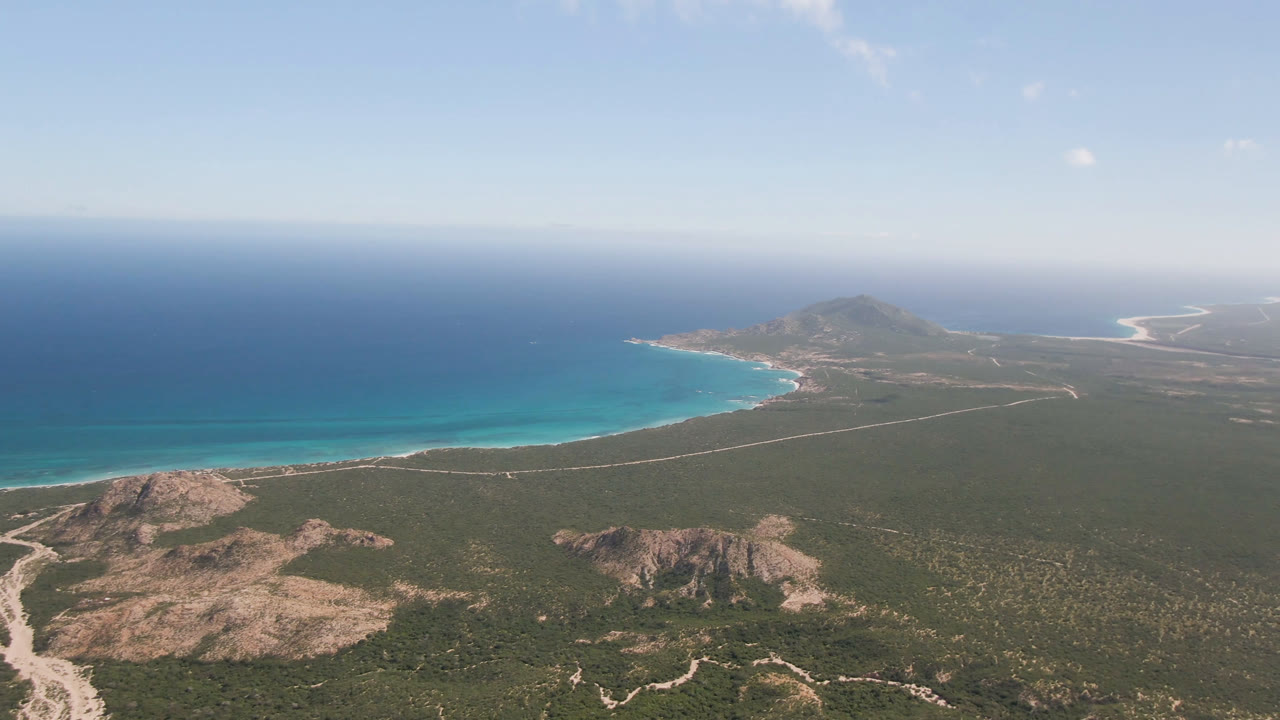 Aerial view over Cabo Pulmo coastline showing more of the curved bay, turquoise water along the sand, low rocky hills below, a distant headland, and no visible people.