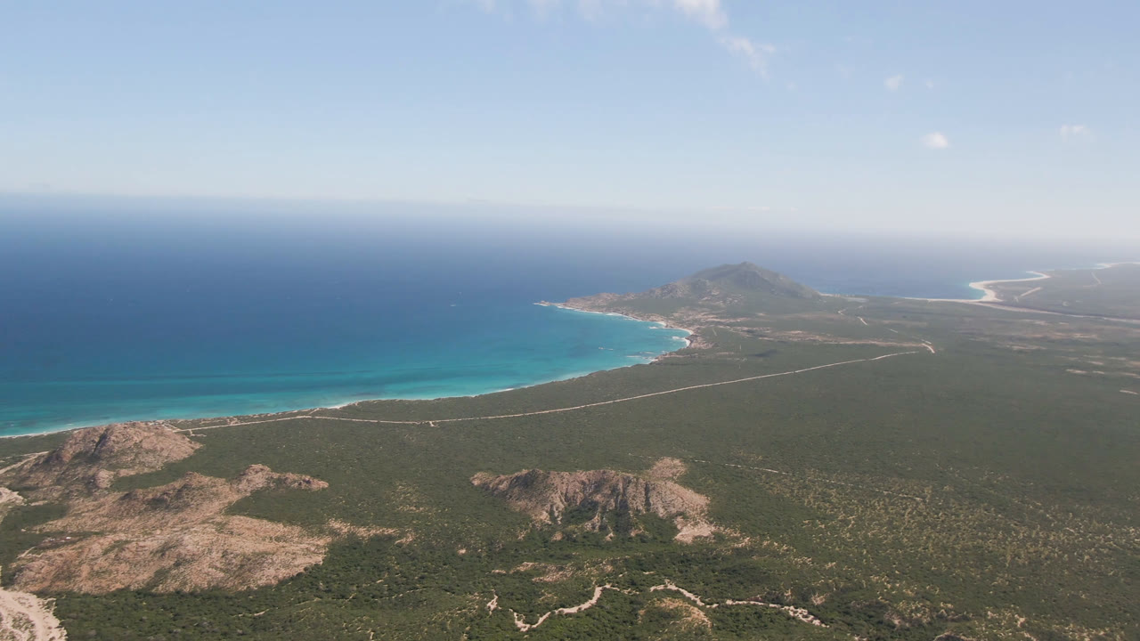 Aerial coastline view with open sea, pale beach, scrubland, and distant landforms.
