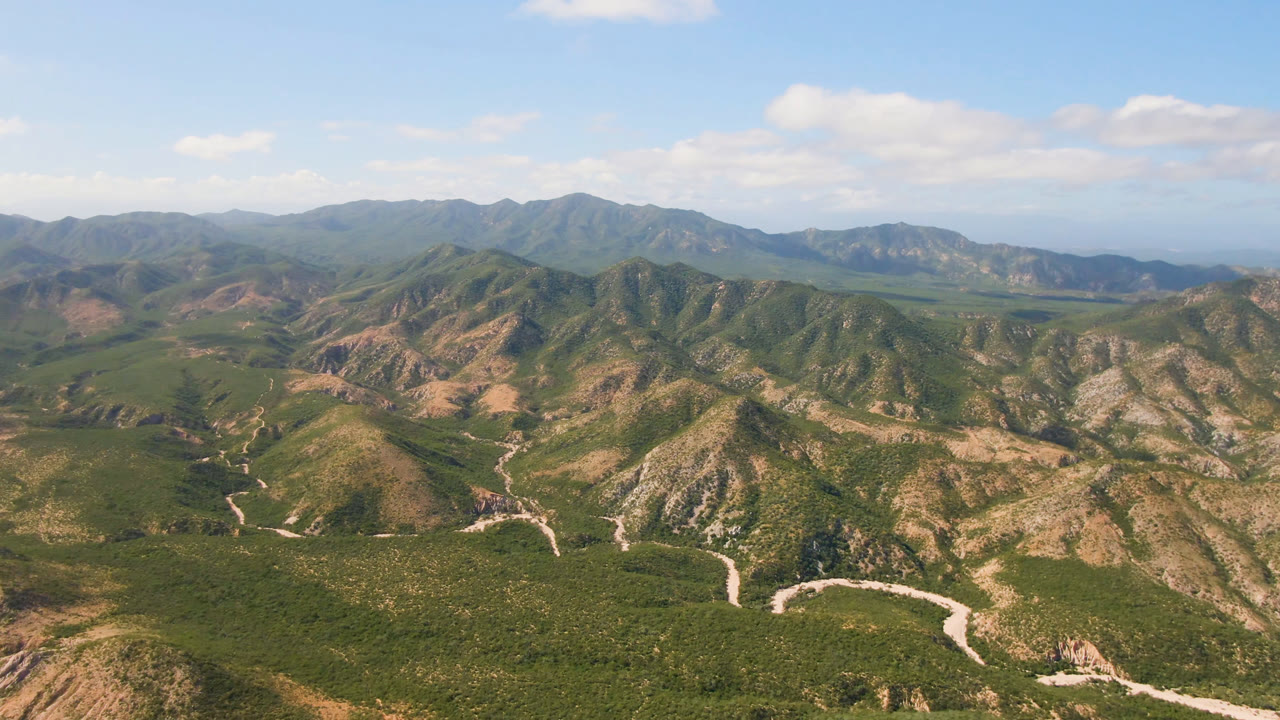 Opening aerial frame over Cabo Pulmo, Baja California Sur, showing rugged green and tan hills, a pale winding road or wash in the valley, distant ridges, soft clouds, and no visible people.