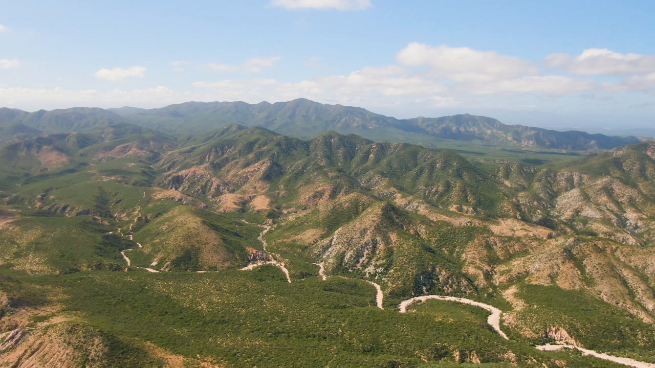 Second frame of the Cabo Pulmo aerial view, with the winding light-colored route more centered among scrub-covered slopes, layered mountains on the horizon, blue sky, and no visible people.