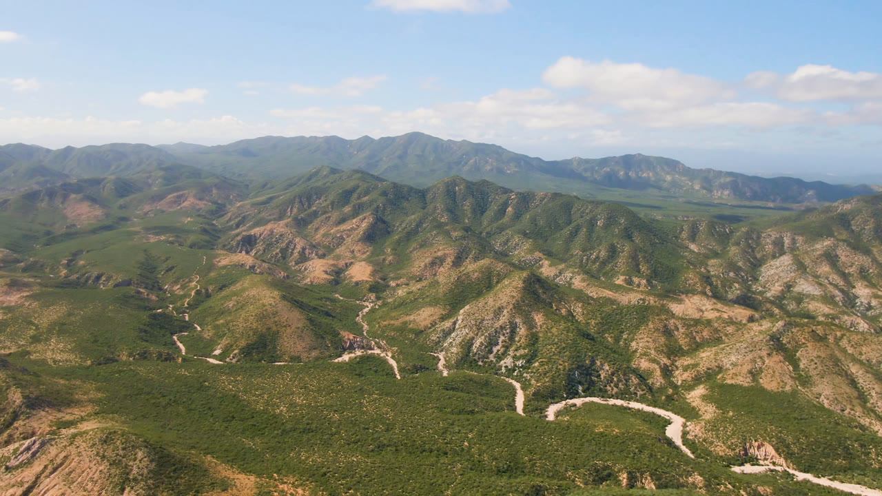 Aerial mountain landscape with ridges, green valley vegetation, and bright clouds overhead.
