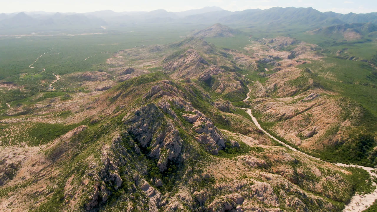 Aerial view in Cabo Pulmo with a rocky central ridge, scattered green vegetation, pale road or wash lines, distant hills, and no visible people.