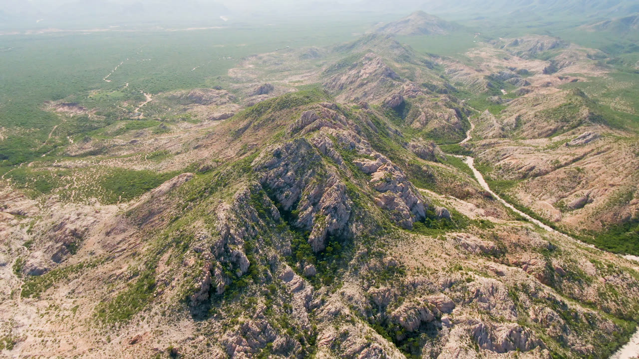 A high aerial frame over rocky mountains and green valley brush in Cabo Pulmo, Baja California Sur, Mexico, with a pale road or wash cutting through the terrain and no visible people.