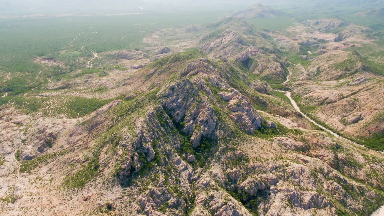 A centered aerial view of the Cabo Pulmo mountain spine, showing rocky slopes, patchy green vegetation, a narrow pale route through the valley, and no visible people.