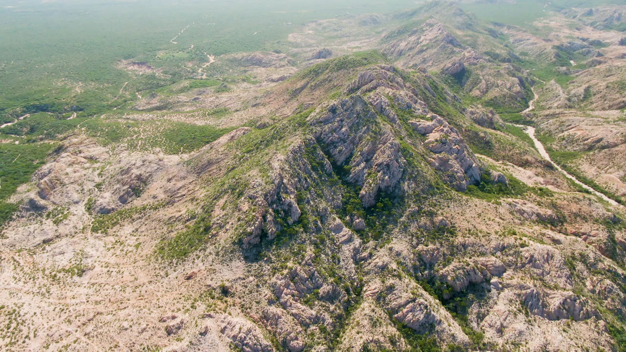High aerial view over rocky hills and green desert scrub in Cabo Pulmo, Baja California Sur, with a pale winding track through the valley and no visible people.