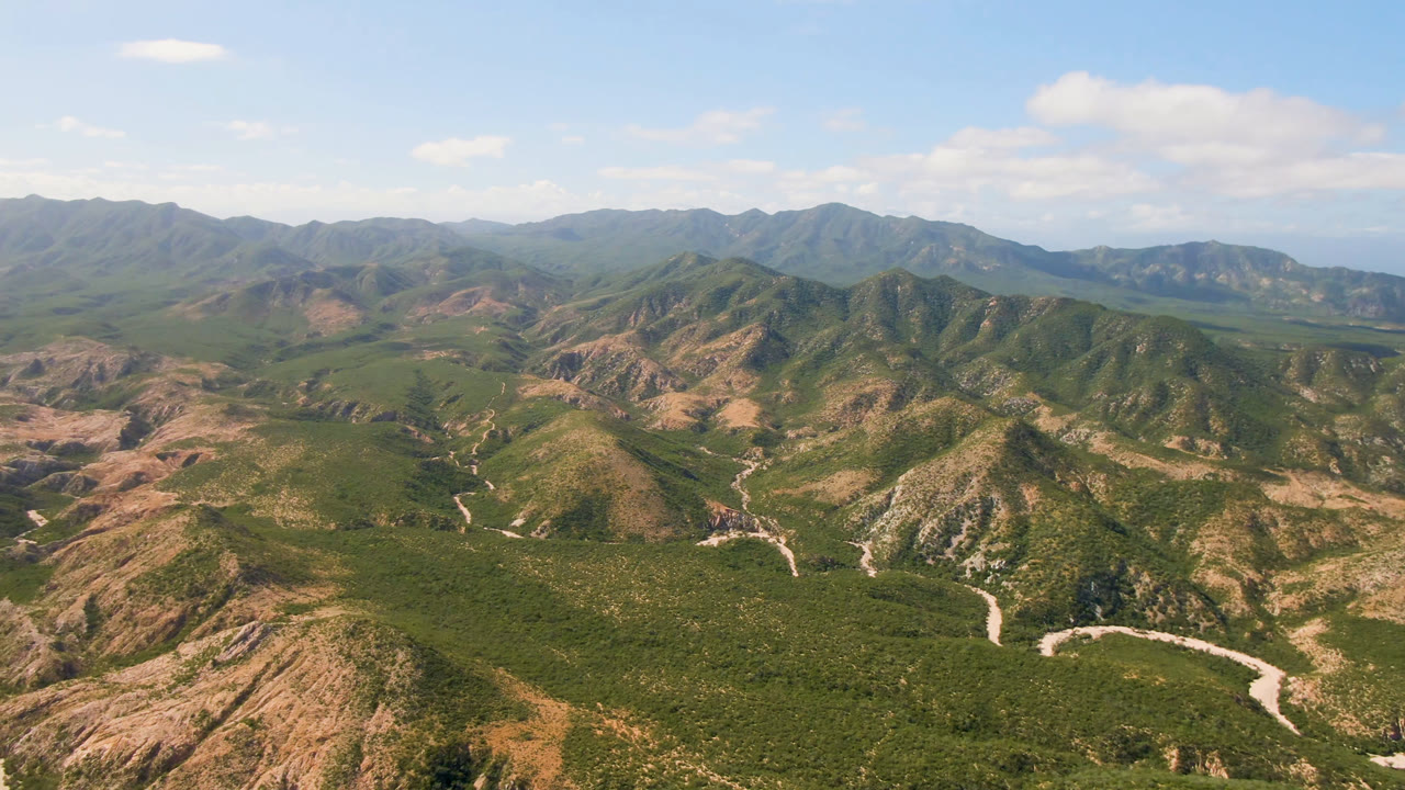Opening aerial frame over Cabo Pulmo mountain ridges, with pale dirt roads crossing the lower valley, distant ranges on the horizon, blue sky, and no visible people.