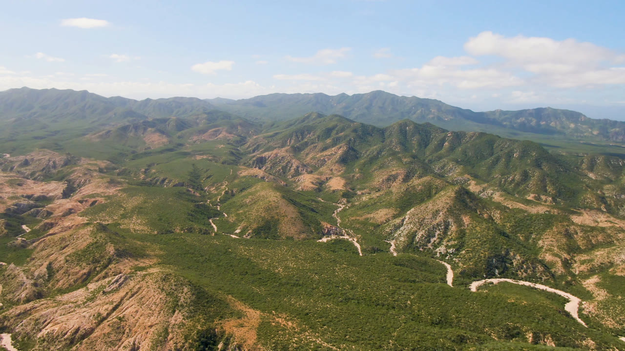 Second aerial frame in Cabo Pulmo showing a broad green valley, tan eroded slopes, a pale road curving near the bottom edge, layered mountains, and no visible people.
