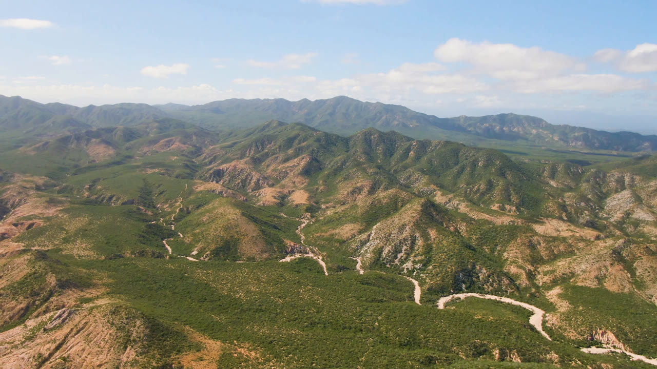 Opening aerial frame over Cabo Pulmo, Baja California Sur, Mexico, showing rugged green and tan mountain ridges, pale winding roads, distant water, scattered clouds, and no visible people.