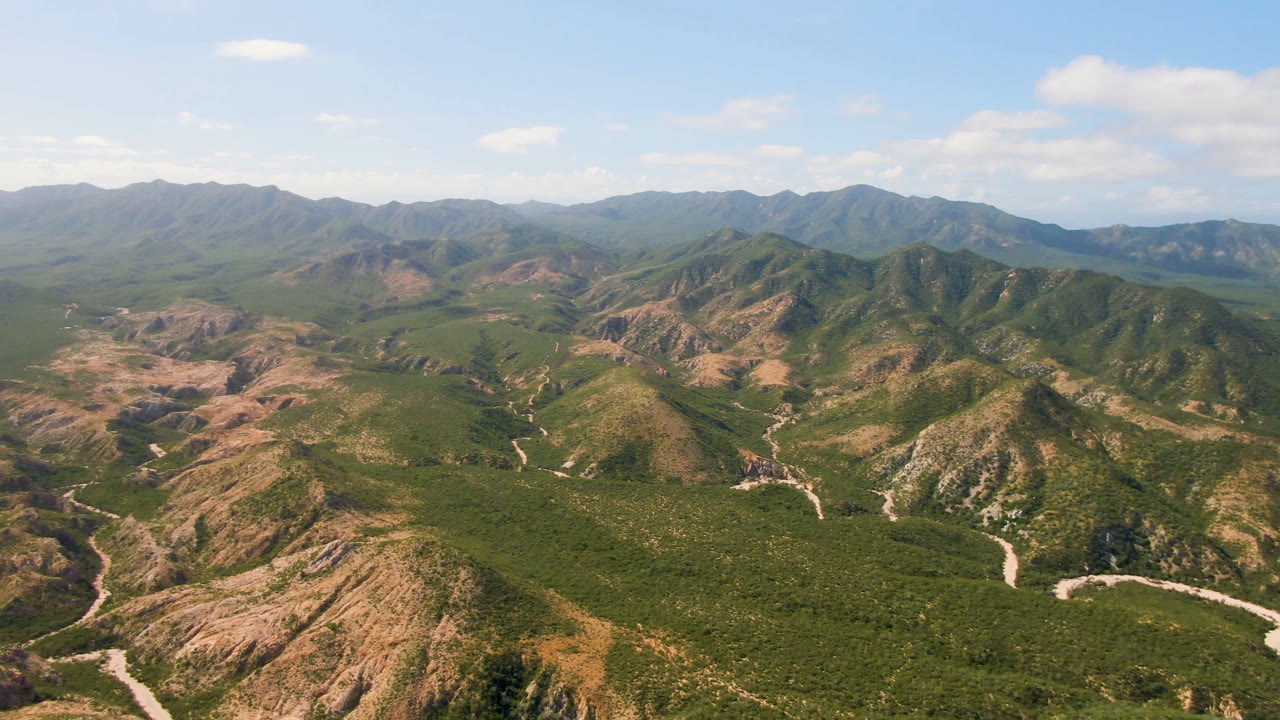Aerial view of green and tan mountains with winding dry washes under blue sky.