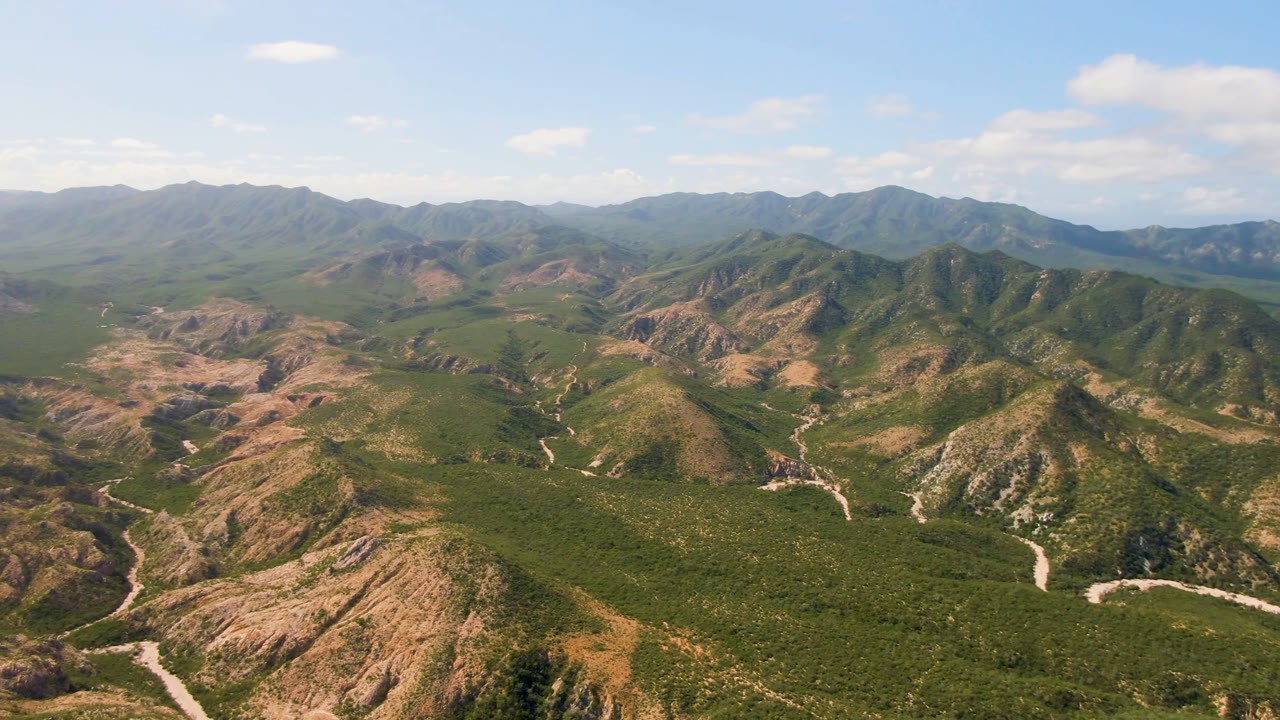 Aerial frame from Cabo Pulmo looking across rugged green and tan hills toward layered mountains, with a pale winding route through the valley and no visible people.