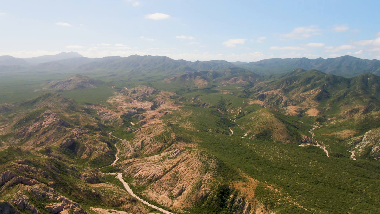 Wide aerial view of Cabo Pulmo mountain ridges and green desert vegetation, with a pale road or wash curving through the valley and no visible people.