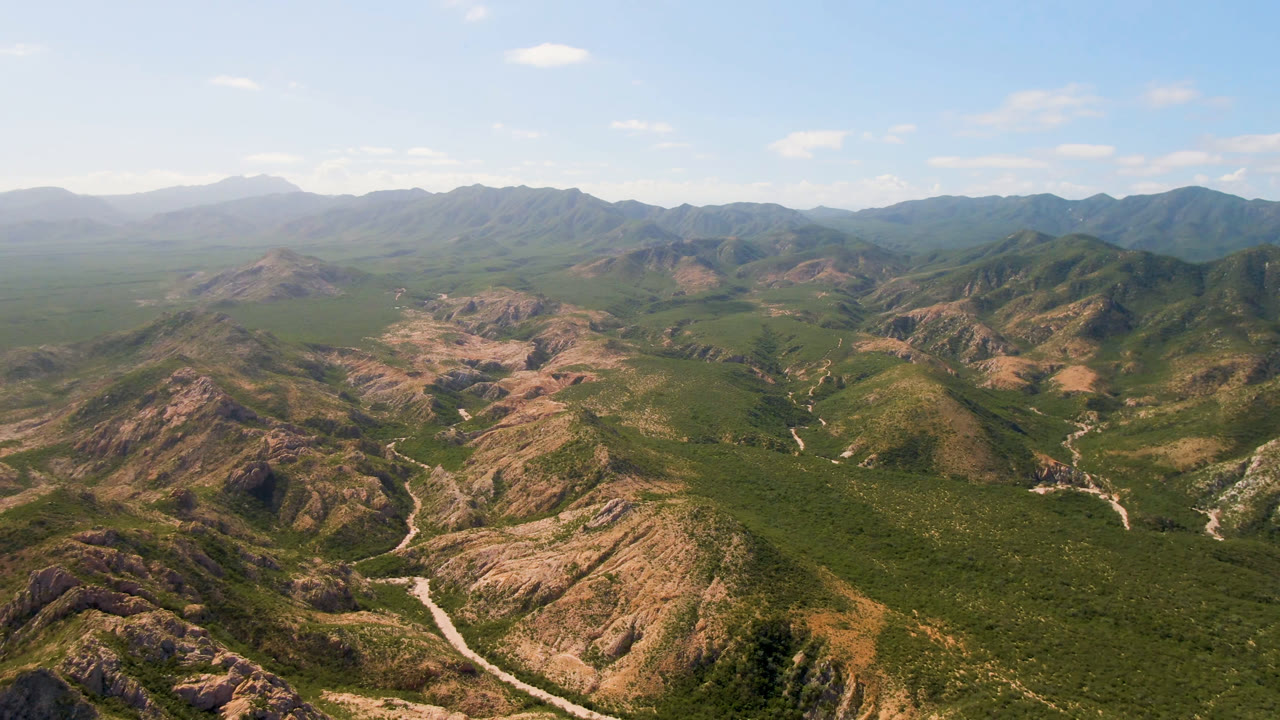 High aerial frame over Cabo Pulmo canyon terrain, showing tan rocky ridges, dense green valley vegetation, distant hazy peaks, and no visible people.