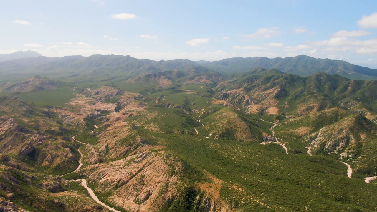Opening aerial frame over Cabo Pulmo, Baja California Sur, Mexico, showing green and tan mountain ridges, a pale winding road through the valley, distant hills, blue sky, and no visible people.